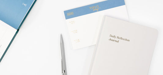 A white daily reflection journal and a top 3 today daily planning pad on a white desk