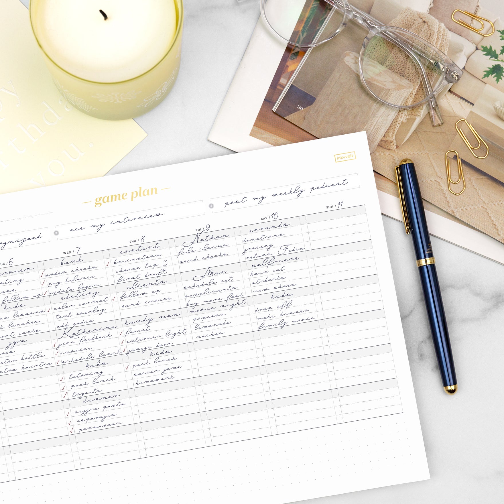 A game plan deskpad filled in with a blue pen, next to a gold candle on a white desktop