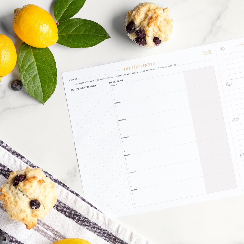An Ink+Volt meal planning pad on a marble counter, next to a berry scone and a yellow lemon with green leaves attached.