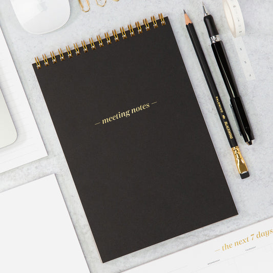 A black meeting notes pad with a black pencil and pen on a white table