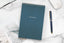 A dark blue gemstone spiral one-on-one pad on a white desk next to a pen