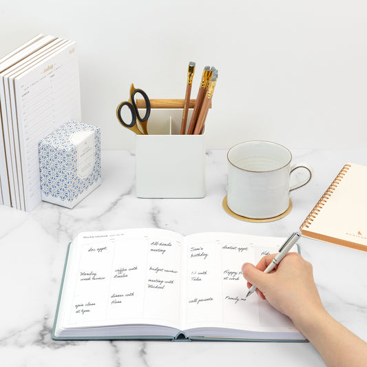 A hand holding a pen writing plans in a light blue planner on an organized desk with blue and white accessories