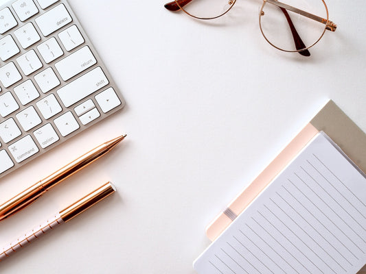 A clean white desk with a keyboard, eyeglasses, paper, and two rose gold pens