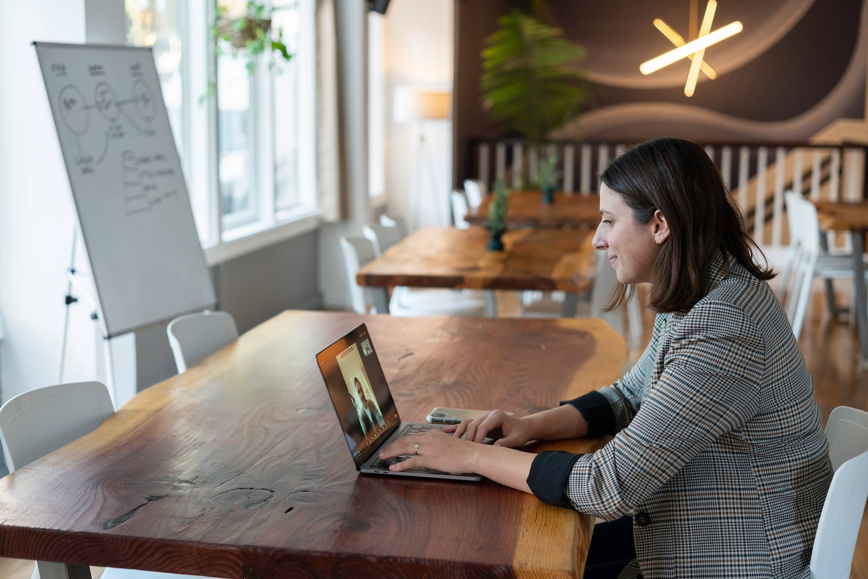 A woman sits at a table in front of a whiteboard, talking to someone on a video call on the computer