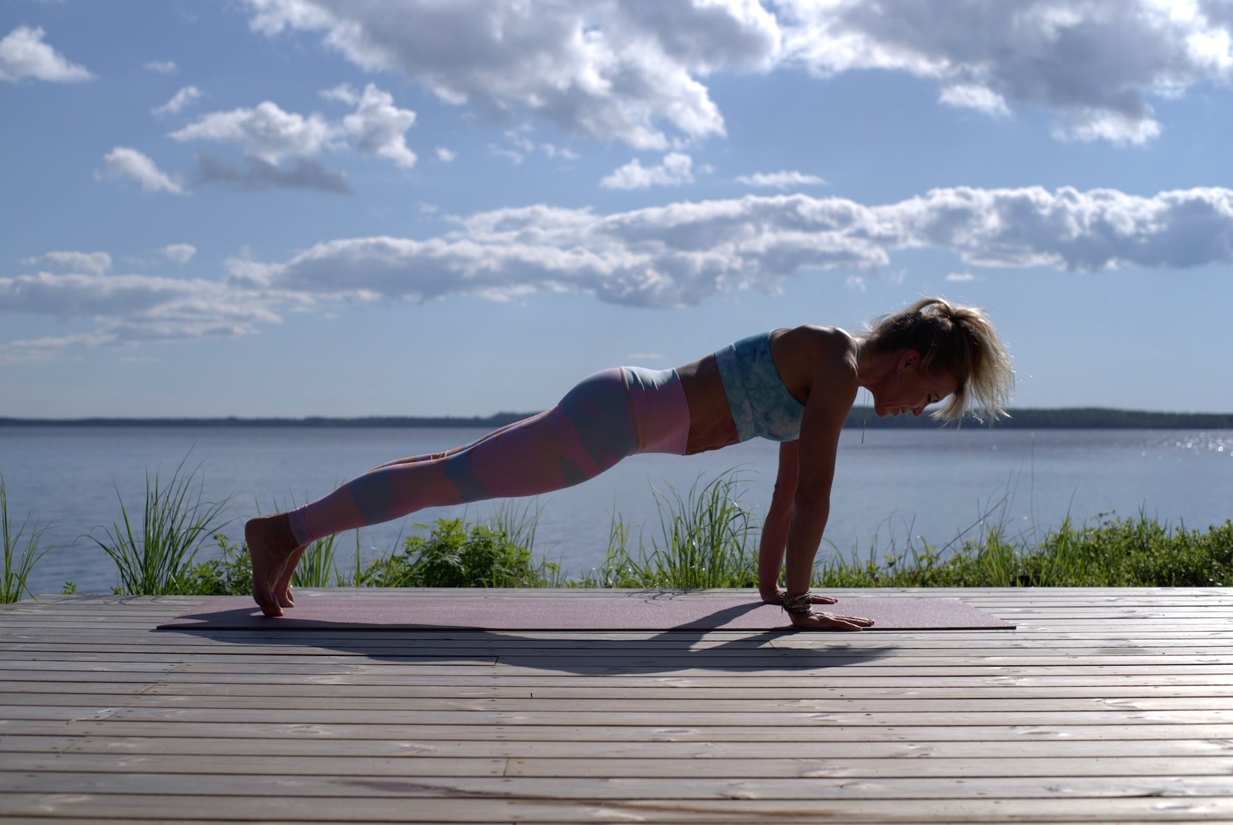 A woman does a plank on a wooden dock in front of a lake and a blue sky