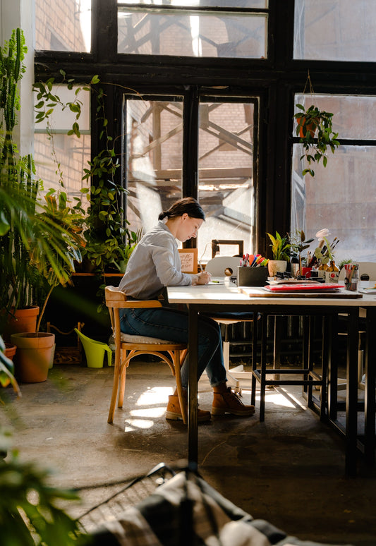 A woman smiles working at a large work table surrounded by lush plants and big windows