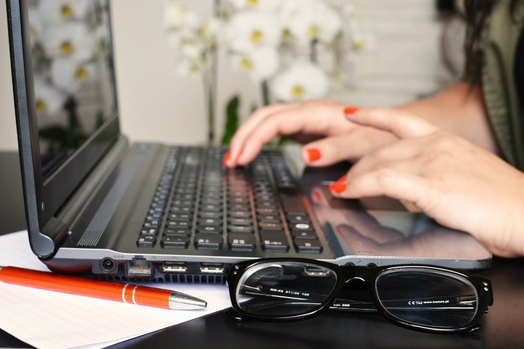 Hands with red nail polish type on a laptop with a set of eyeglasses next to the computer