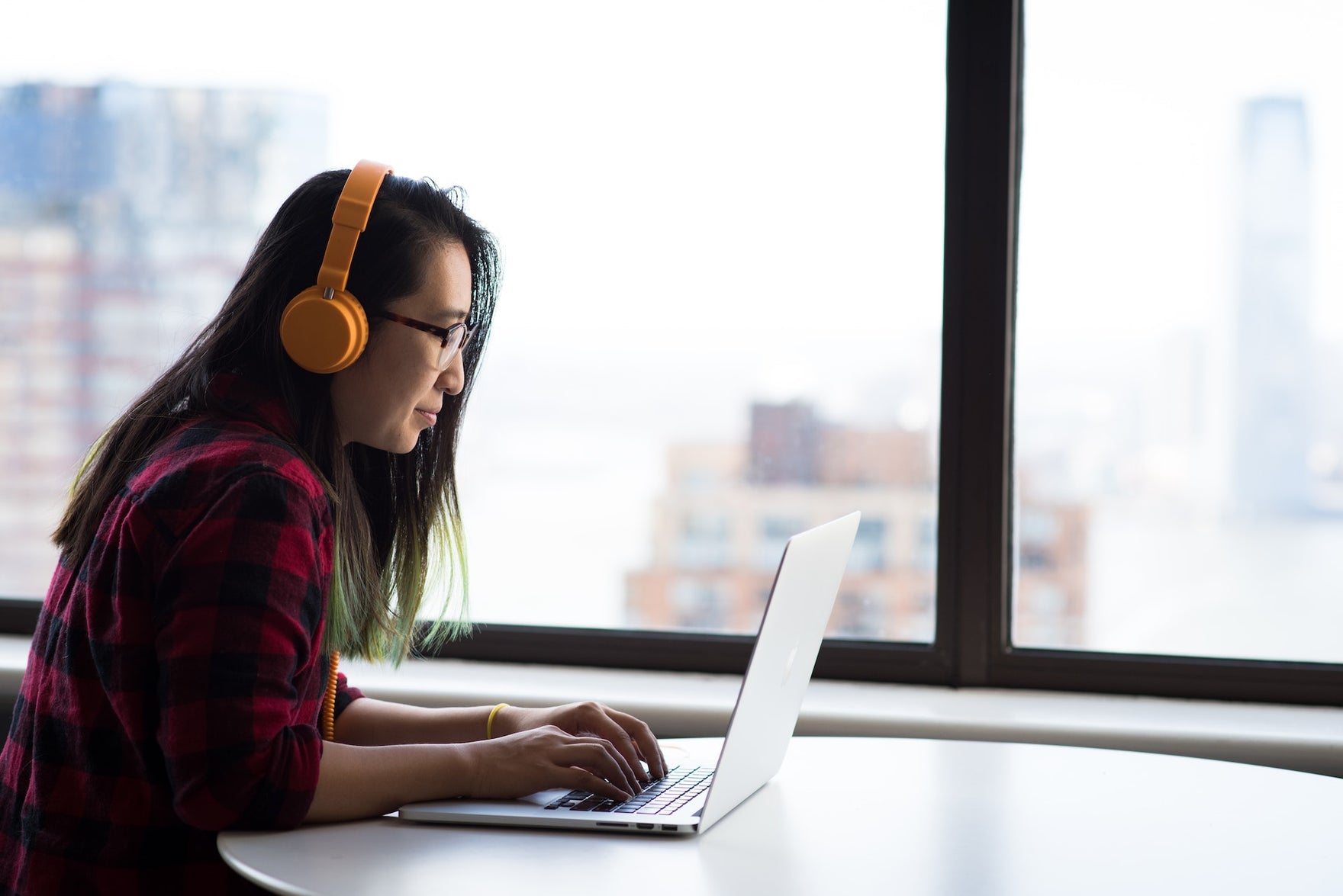 A woman with headphones works at a laptop in front of a big window