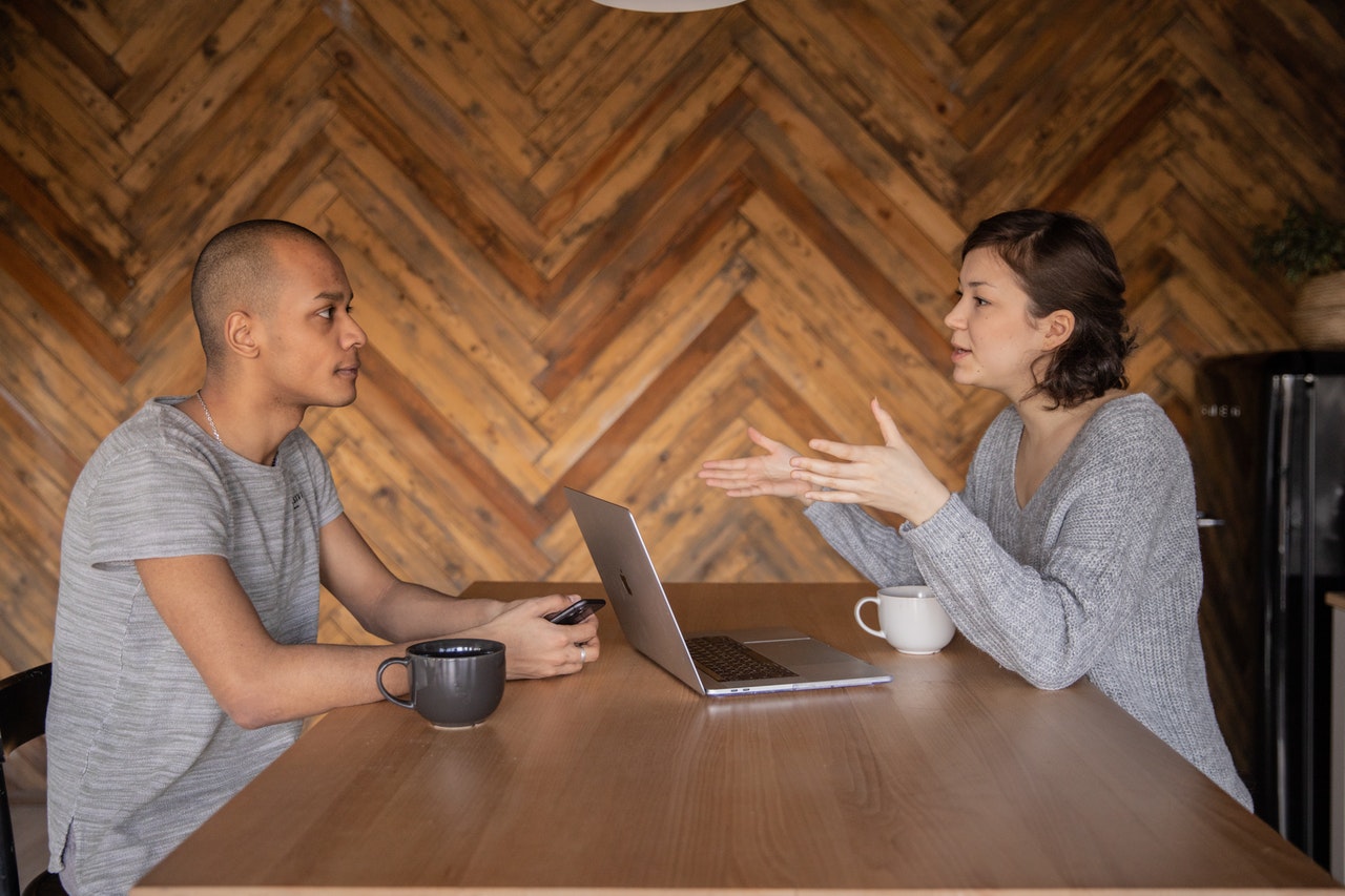 A man and a woman sitting and talking at a table with a laptop.