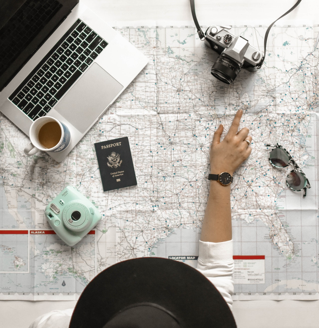 An assortment of travel items on a map - camera, sunglasses, passport, and a woman's hand pointing at the map.