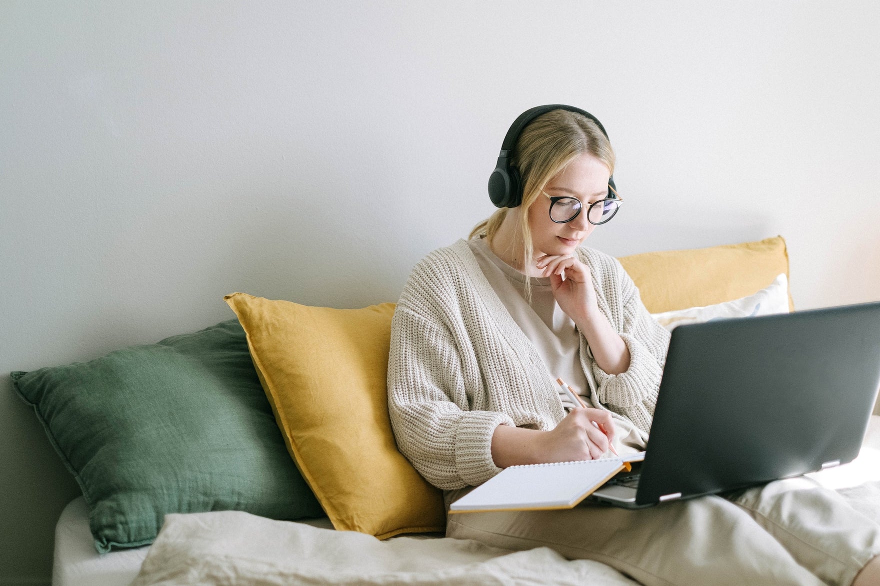 A blond woman sits on a couch working on a laptop and wearing headphones