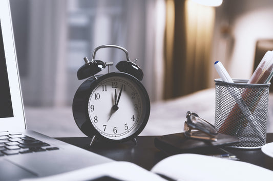 A black alarm clock sits on a desk next to a laptop and a wire pencil cup