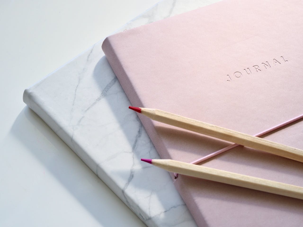 A pink notebook on top of a white notebook, with two colored pencils on top