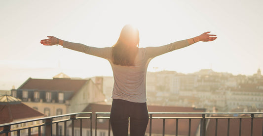 A woman stands on a balcony with her arms outstretched toward the sun