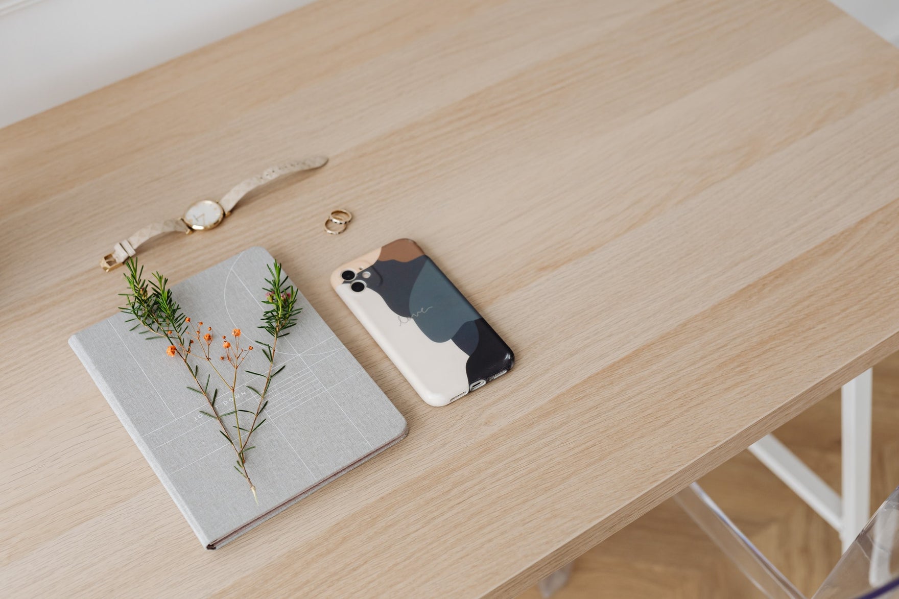 A grey notebook with sprigs of greenery on a light wooden desk, next to a watch and a phone