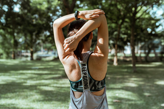 A woman with brown hair stretches her arms overhead in a park