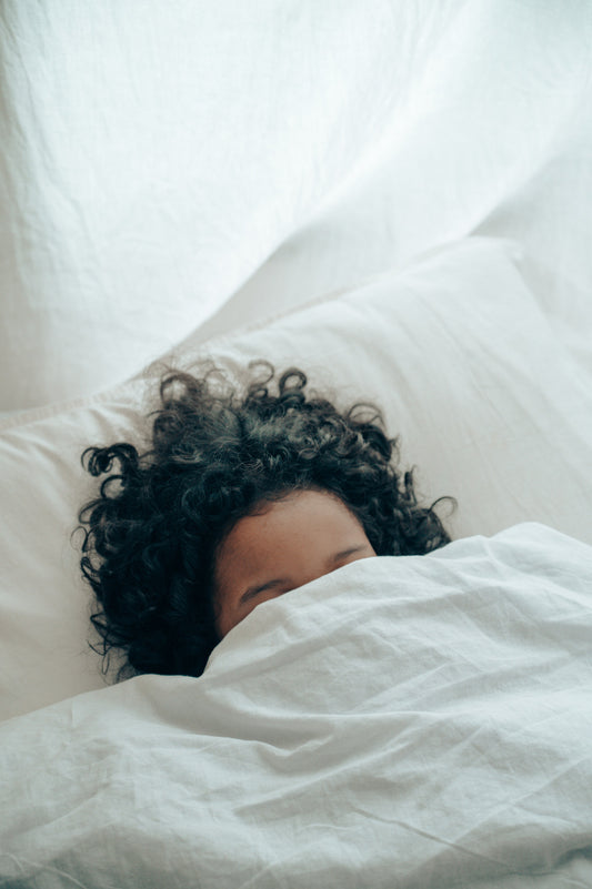 how to get more deep sleep: a woman with brown wavy hair puts her head under a white blanket