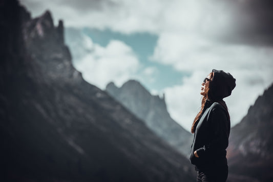 A woman in a black sweater and sunglasses stands looking up in front of a mountain range.