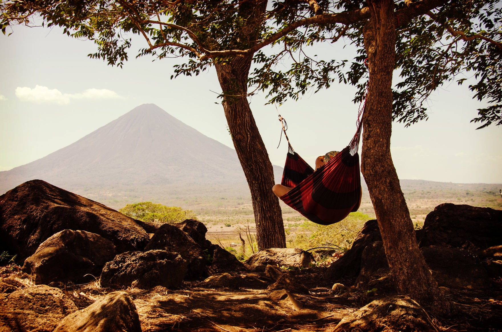 A person relaxes in a hammock between two trees in front of a desert mountain.