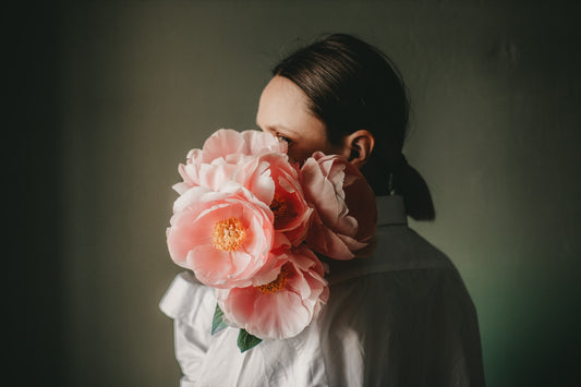 A white woman holds a bouquet of big pink flowers over her shoulder, covering her face.