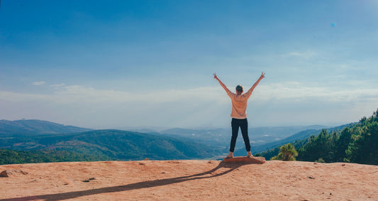 A person stands on a cliff with arms raised in the air in front of a blue sky