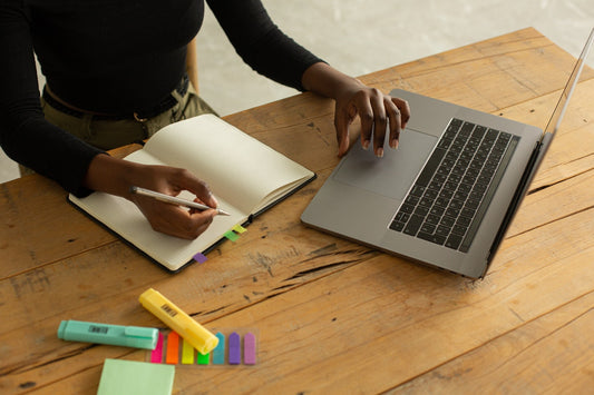 A woman works at a desk while writing in a notebook, next to a laptop and highlighters.