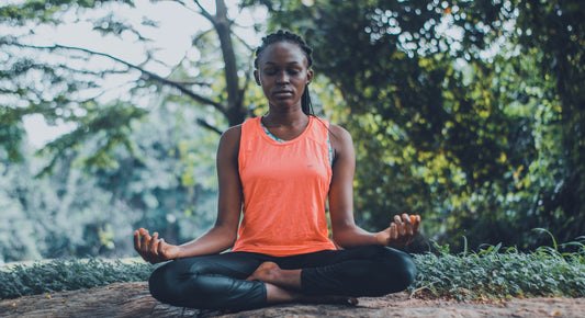 A woman sits in lotus pose outside in front of a leafy background