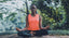 A woman sits in lotus pose outside in front of a leafy background