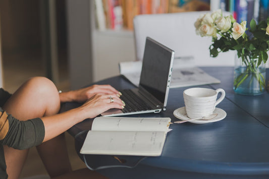 A woman sitting comfortably at a table with a laptop, a notebook, a cup of tea, and a vase of roses.