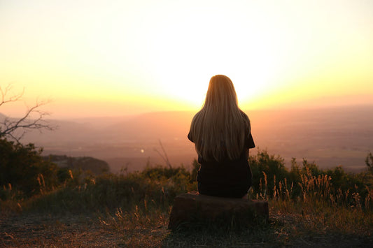 A woman sits outside in front of a sunset over hills