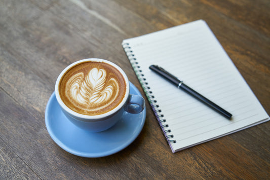 A latte next to a notebook and pen on a wood table.