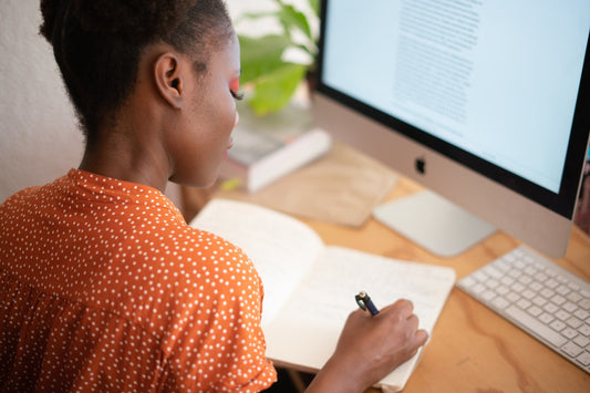 A woman sits in front of a computer while writing in a notebook