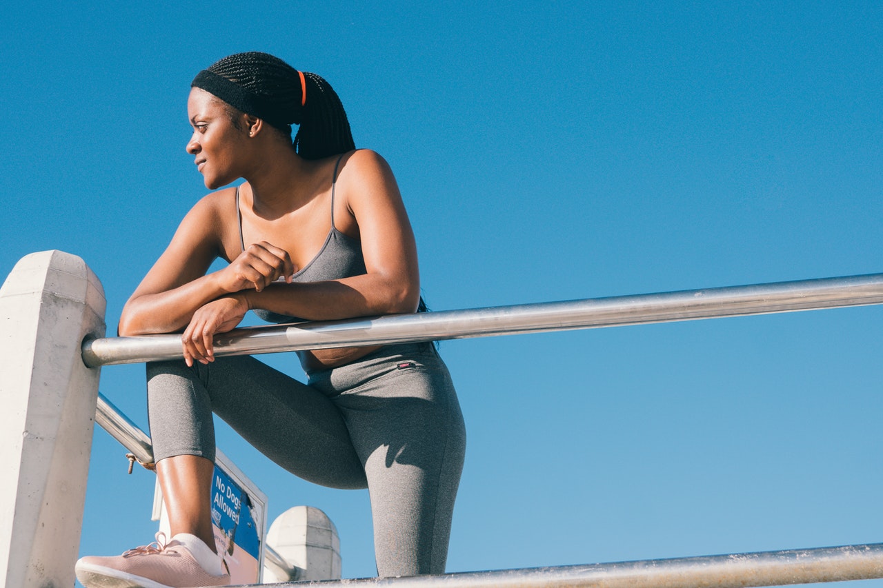 A woman in grey workout gear leans on a railing in front of a bright blue sky.