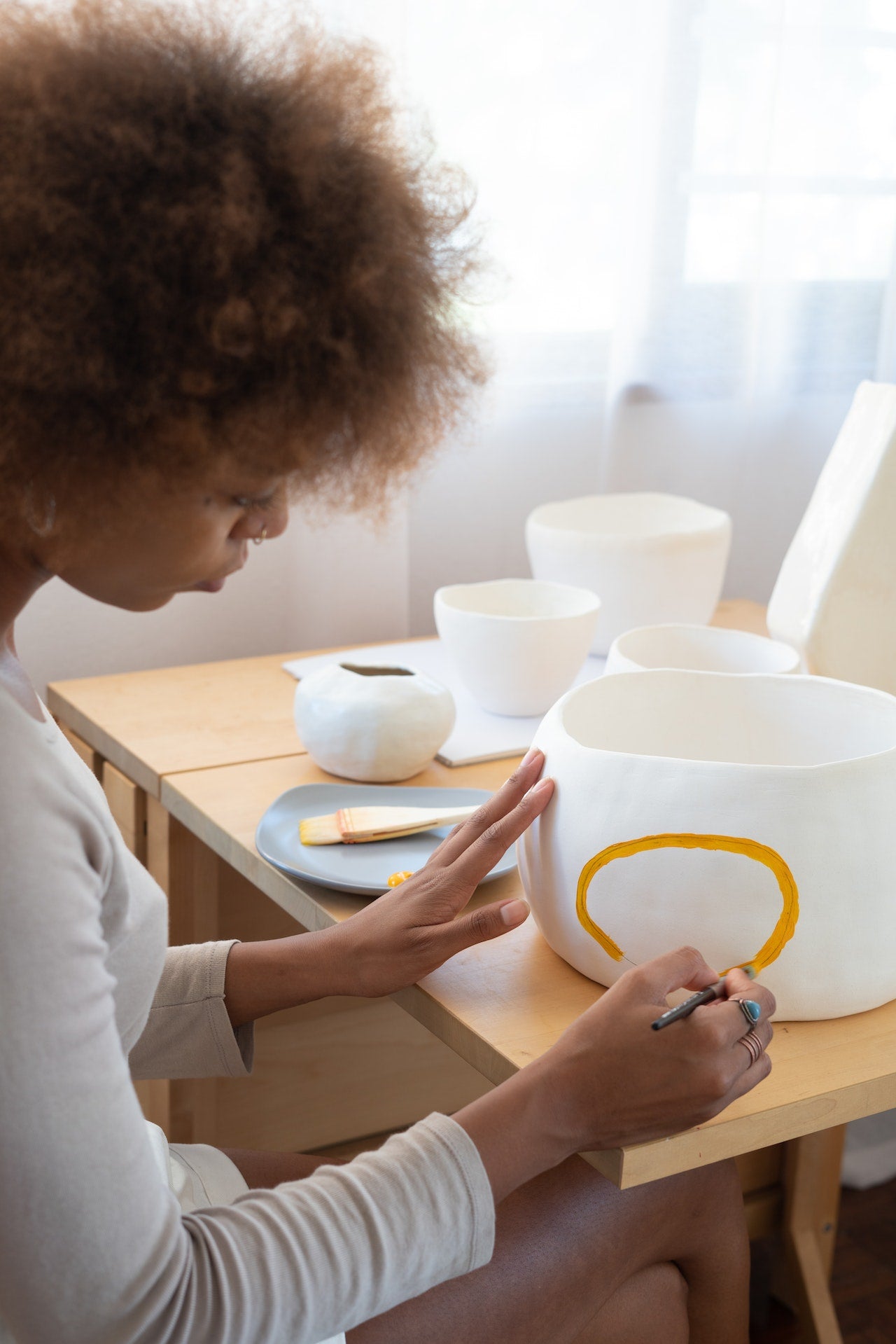 A woman sits at a table painting a gold line on a white ceramic pot