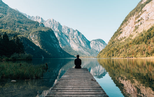 A person sits at the end of a dock on a lake in front of mountains.