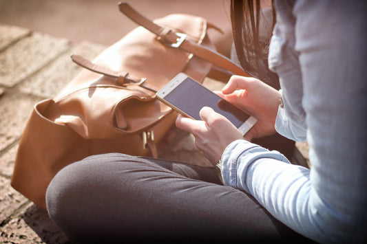 A woman sits outside looking at her phone, with a leather bag sitting in front of her.