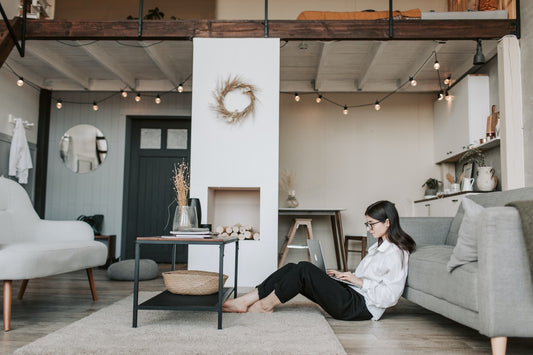 A woman sits on the floor of a neutrally styled apartment working at a laptop on her lap.