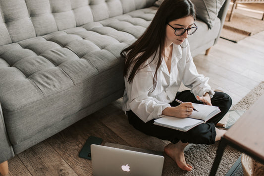 A woman in glasses sitting in front of a grey couch writes in a notebook, next to an open laptop on the floor