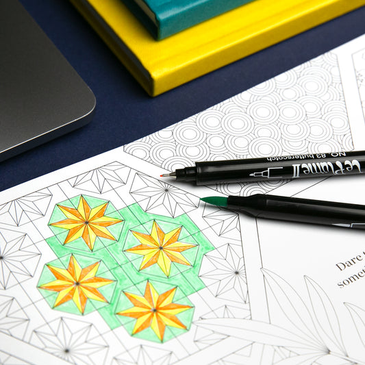 Coloring book with geometric designs, markers, and a laptop on a blue surface