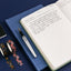 Open notebook with handwritten text on a blue surface, accompanied by a pen and floral tape.