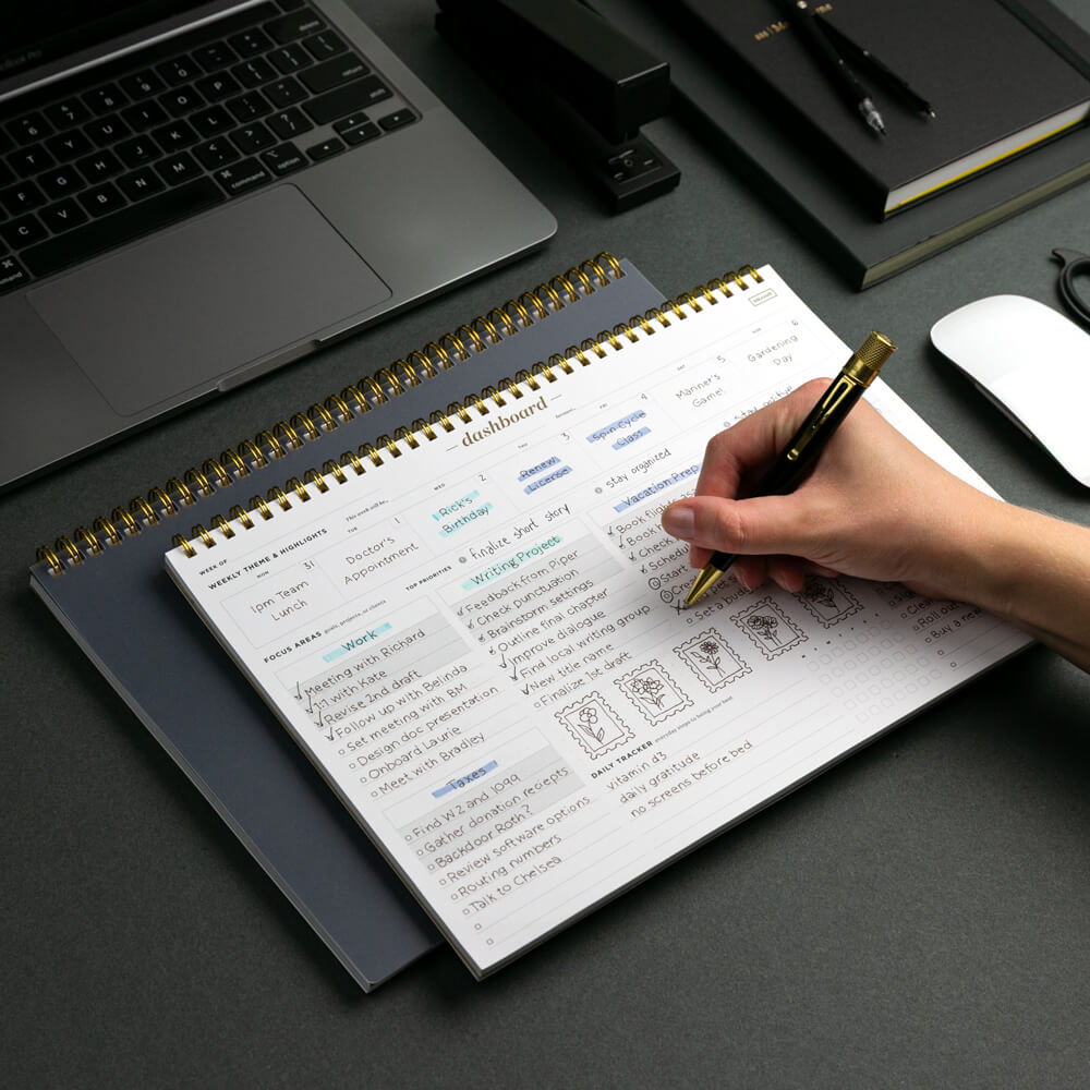Person writing in a planner with a laptop and office supplies on a desk