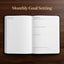 An open planner or notebook with monthly goal-setting pages displayed on a dark wood background, with the text 'Monthly Goal Setting' above it.