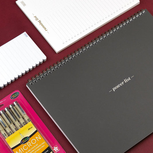 Gray notebook with 'power list' on cover, surrounded by white notebooks and pink Micron pen set on a red surface.
