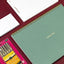 Green spiral-bound notebook with 'power list' text, white notebook, and pink micron pen set on a red surface.