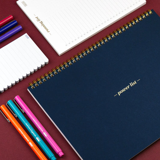 Blue notebook with 'power list' on a red surface with colorful pens.